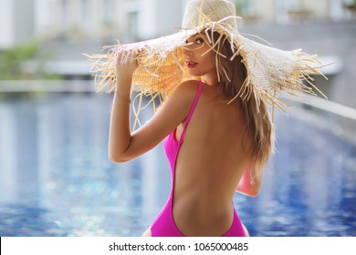 Summer lifestyle fashion portrait of young stunning blonde woman sitting half-turn near the pool. Sunbathing, enjoying life. Wearing stylish pink swimsuit, beautiful straw hat with wide brim. Close up