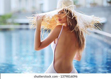 Summer lifestyle fashion portrait of young stunning blonde woman sitting half-turn near the pool. Sunbathing, enjoying life. Wearing stylish white swimsuit, beautiful straw hat with wide brim. Closeup
