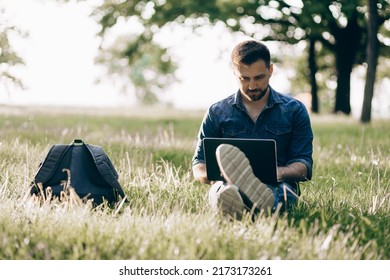 Summer lifestyle handsome bearded guy european businessman sitting on green grass in blue denim jacket holding macbook in hands and typing