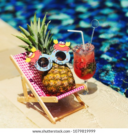 Summer lifestyle image of young pretty girl-pineapple lying on sun chaise near the turquoise swimming pool. Wearing funny sunglasses. Drinking fresh juice. Tropical summer vacation concept. Sunbathing