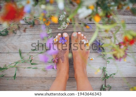 Summer lifestyle portrait of a attractive girl's feet with buds of wildflowers between toes. Staying on wooden terrace surrounded by flowers. Enjoying life, nature. Feet in focus, flowers in blur. 