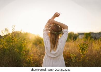 Summer lifestyle portrait of beautiful young woman in a wreath of wild flowers. Standing back in the flower field, hands to the side. Romantic mood. Nature lover