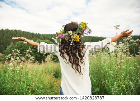 Summer lifestyle portrait of beautiful young brunnete woman with perfect long curly hair in a wreath of wild flowers. Standing back in the flower field, hands to the side. Romantic mood. Nature lover 