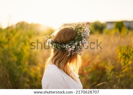 Summer lifestyle portrait of beautiful young woman in a wreath of wild flowers. Standing back in the flower field, hands to the side. Romantic mood. Nature lover