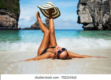 Summer lifestyle portrait of happy pretty young woman with tanned sexy body. Enjoying life and lying in the clear sea water on the beach of the tropical island. Stylish wide brimmed hat. Leg up 