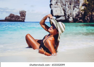 Summer lifestyle portrait of pretty young suntanned woman. Enjoying life, smiling and lying on the beach of the tropical island. Wearing stylish bikini, sunglasses and wide brimmed hat with stripes.
