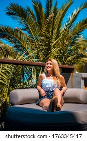 Summer Lifestyle Of A Young Blonde Caucasian Woman In A White Flowery Top Enjoying The Summer On A Terrace With Palm Trees. Sitting On A Sofa On A Terrace Sunbathing