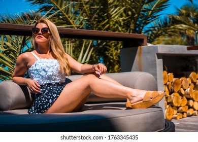 Summer Lifestyle Of A Young Blonde Caucasian Woman In A White Flowery Top Enjoying The Summer On A Terrace With Palm Trees. Sitting On A Sofa On A Terrace