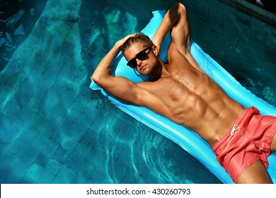 Summer Man Body Sun Skin Care. Beautiful Model With Sexy Body In Swimwear Tanning, Floating On Mattress In Swimming Pool Water. Fitness Male With Healthy Tan Relaxing At Relax Spa Resort. Summertime