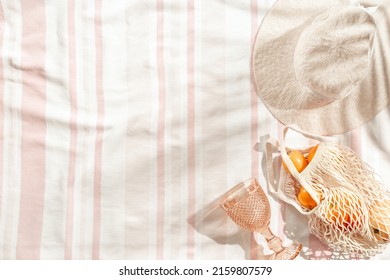 Summer minimal flatlay with wide-brimmed hat, colored wine glass, orange fruits in mesh bag on stripes beach towel as background, sun shadows. Summer pastel colored aesthetic photo, copy space