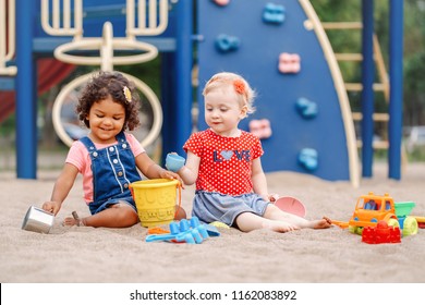 Summer Mood. Two Cute Caucasian And Hispanic Latin Babies Children Sitting In Sandbox Playing With Plastic Colorful Toys. Little Girls Friends Having Fun Together On Playground.