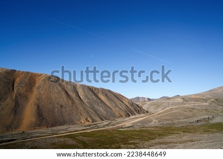 Summer mountain arctic landscape. View of the rocky hills. In the blue sky, the moon and a flying plane. Road through the tundra and mountains. Northern nature. Travel and hiking in the Arctic. July.