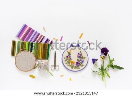 Summer needlework. Hand embroidery with satin ribbons of blooming pansies in round hoop, sets of multi-colored ribbons, accessories for embroidery on white background. Flat lay, copy space, close-up