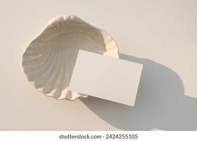 Summer neutral wedding stationery. Blank greeting, RSVP or business card mockup in sunlight. Invitation, template. Beige sea shell plate. White table background. Elegant composition, flatlay, top view