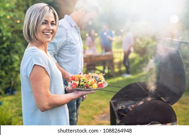 In summer. A nice couple prepares a bbq to welcome friends in the garden. She's holding a plate of grilled skewers - Powered by Shutterstock