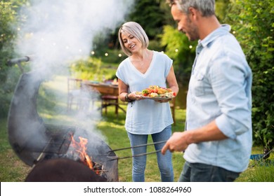 In summer. A nice couple prepares a bbq to welcome friends in the garden. She's holding a plate of grilled skewers - Powered by Shutterstock