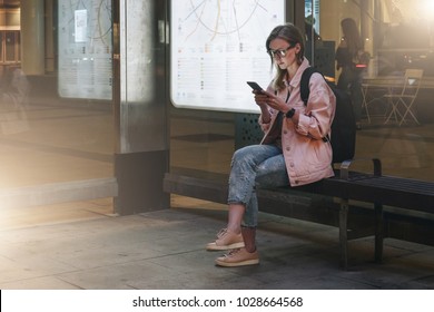 Summer night. Young woman in eyeglasses with backpack is sitting at bus stop and is using smartphone. Hipster girl writes message, chatting, browsing internet, checking email. Social media, network.