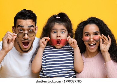 Summer Offer. Funny Excited Arab Mother, Father And Daughter Wearing Colorful Modern Sunglasses, Amazed Middle Eastern Parents Looking At Camera With Opened Mouth, Posing Over Yellow Background