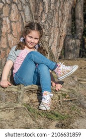 In summer, on a bright sunny day, a small beautiful girl in sneakers sits under a pine tree.