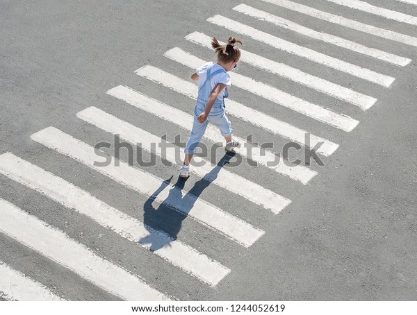 In the summer on the street at the\
pedestrian crossing kid girl in fashion clothes cross the road.\
From top view. Behind. Shadow at zebra\
crossing