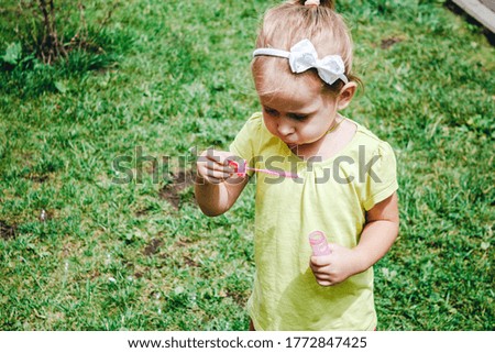 Summer outdoor activities for children. A little blonde girl in a yellow t-shirt blows soap bubbles against the background of green bright grass in the courtyard with a blur of focus.