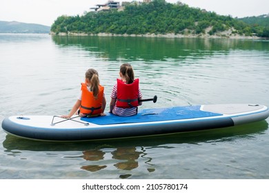 Summer outdoor activities with kids concept. Two girls sitting on inflatable stand up paddle board. Children enjoying peaceful paddling during family  local getaway. Children learning paddleboarding