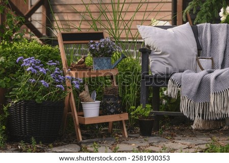 Summer outdoor backyard decorations. Rustic flower stand with potted plants, wooden bench with pillows and blanket. Cozy garden relax area.