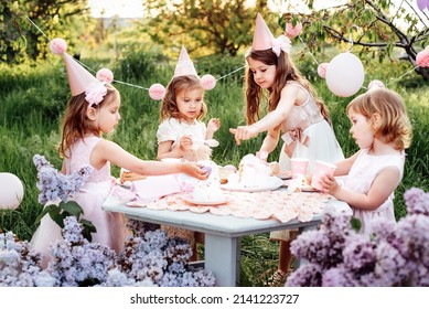 Summer Outdoor Kids Birthday Party. Group Of Happy Children Celebrating Birthday In Park. Children Blow Candles On Birthday Cake. Kids Party Lilac Pink Pastel Decoration And Food. Presents And Sweets.