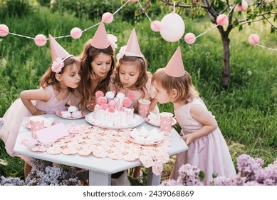 Summer outdoor kids birthday party. Group of happy Children celebrating birthday in park. Children blow candles on birthday cake. Kids party pink pastel decoration and food. Presents and sweets. - Powered by Shutterstock