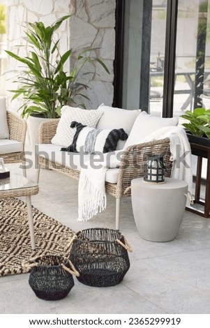 Summer Outdoor Patio on a Balcony of a Modern Luxurious House, Featuring a Wicker Sofa with Beige Cushions, Concrete Side Table, Tempered Glass Table, and Furniture Cover, on a Sunny Day.