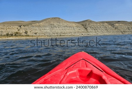 Summer outdoor recreation watersport image of the bow of a red kayak floating on the water of a river with a tall sandstone cliff on the riverbank.