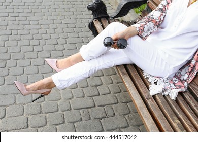 Summer outfit, street fashion look. Young tanned beautiful woman in white pants and light floral pink patent in leather high-heeled shoes. Stylish girl sitting on a bench in the city. 