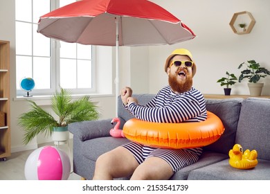 Summer party at home. Funny chubby man having fun sitting on sofa in living room with inflatable swimming circle. Humorous man has absurd vacation under beach umbrella on improvised home beach. - Shutterstock ID 2153541739