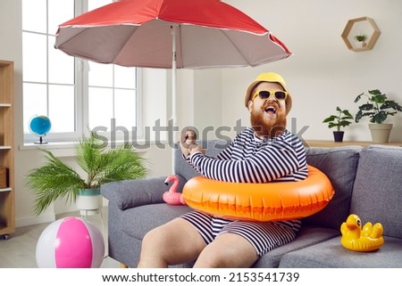 Summer party at home. Funny chubby man having fun sitting on sofa in living room with inflatable swimming circle. Humorous man has absurd vacation under beach umbrella on improvised home beach.