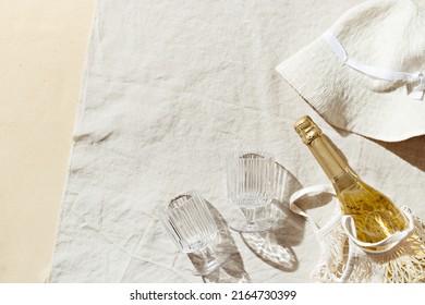 Summer Pastel Background With Sun Hat, Wine Glasses, Bottle Sparkling Wine In Mesh Bag On Cotton Beach Towel As Background, Sunlight Shadows. Concept Of Summer Hot Day On Beach, Picnic With Hard Sun