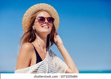 Summer photo of a pretty woman in sunglasses, listening to music in earphones, on smartphone, outdoors. Dressed in stylish clothes and hat. - Powered by Shutterstock