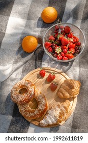 Summer Picnic With A Basket Of Croissants And Bagels On Rustic Wooden Desk.  French Tasty Picnic Top View, Copy Space. Strawberries, Oranges, Cherries In Pile. Beach Grey Black Blanket.Camping Snack
