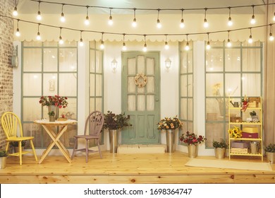 Summer Porch With Garlands Of Light Bulbs. Terrace Of Summer House Design Of Photo Studio. Patio Verandah With Flowers In Metal Buckets And Wooden Chairs And Table. Street Garland With Light Bulb