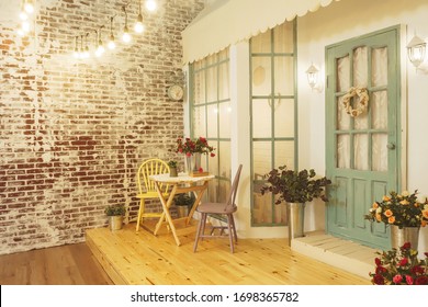 Summer Porch With Garlands Of Light Bulbs. Terrace Of The Summer House Design Of The Photo Studio. A Verandah Patio With Flowers Roses In Metal Buckets And Wooden Chairs And A Table.