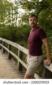 Summer Portrait Of A Caucasian Man In His 40s. He Is Standing On A Wooden Boardwalk At Mer Bleue Bog Located In Ottawa, Canada. He Is Wearing A  Burgundy Shirt And Khaki Shorts. He Is Canadian.