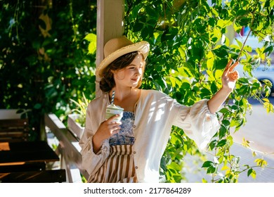 Summer Portrait Of Cute Lovely Teenager Girl In Straw Hat Relaxing In Shadow On Terrace With Curly Grapes On A Hot Day Drinking Cold Drink From A Cup. Summer Relaxing Lifestyle. Village Lifestyle.