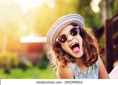 Summer Portrait Of Happy Kid Girl On Vacation In Sunglasses And Hat, Laugh And Showing Tongue. 