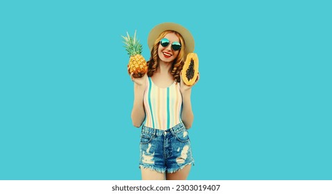Summer portrait of happy smiling young woman with pineapple and papaya, fresh tropical juicy fruits, wearing straw hat, sunglasses on blue background - Shutterstock ID 2303019407