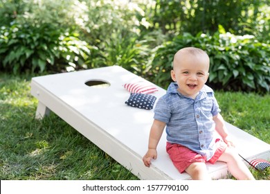 summer portrait of little boy sitting on corn hole game in backyard on 4th of July