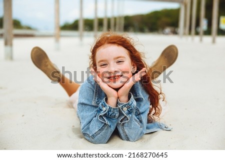 Summer portrait of a little girl with red hair lying on the city beach.