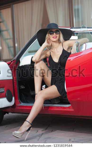 Summer portrait of stylish blonde vintage woman\
with long legs posing near red retro car. Fashionable attractive\
fair hair female near a red vintage vehicle. Sunny bright colors,\
outdoors shot.