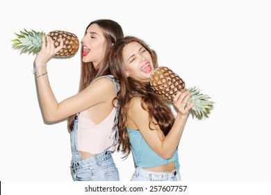 Summer Portrait Of Two Pretty Brunette Girl Friends Having Fun With Pineapple. Singing With Closed Eyes And Smiling. Casual Style, Bright Makeup, Pink Lips. White Background, Not Isolated. Inside