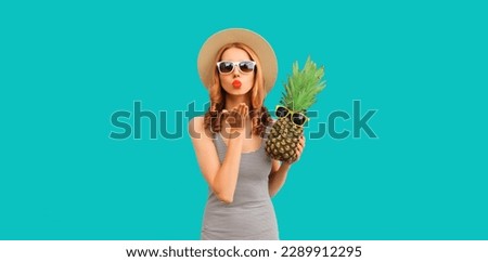 Summer portrait of young woman blowing her lips sending air kiss with pineapple wearing straw hat, sunglasses posing on blue background