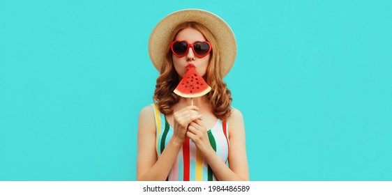 Sommerportrait einer jungen Frau, die eine eiscremeförmige Wassermelone mit einem Strohhut saugt, eine rote, herzförmige Sonnenbrille auf blauem Hintergrund