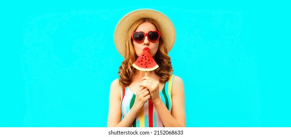 Summer portrait of young woman with juicy lollipop or ice cream shaped slice of watermelon wearing straw hat, red heart shaped sunglasses on blue background, blank copy space for advertising text